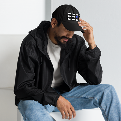 Hombre sentado con Gorra Negra VOOM — The Pattern Code y chaqueta negra posando con gesto pensativo hacia abajo.